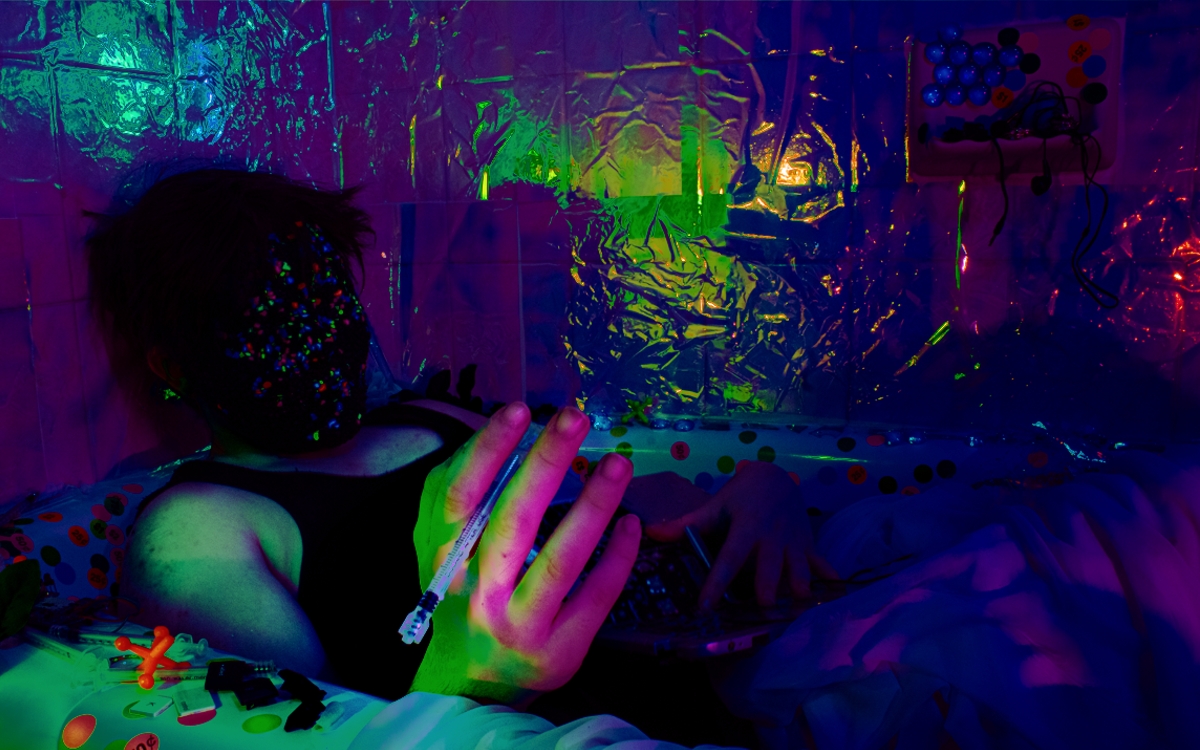 a digital photo of a man in a bathtub wearing a black tank top and a mask covered in neon and black aquarium gravel. he has a syringe without a needle in his hand and a mangled laptop in his lap. the bathtub is covered in colorful neon price tag stickers, keyboard keys, and other random objects. the wall behind is covered in reflective purple and green cellophane.